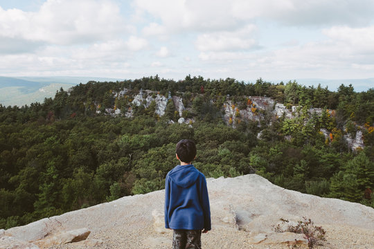 Boy Stands Near Cliff Edge Looking Out At Tree Covered Mountains