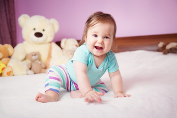 Cute smiling little baby girl sitting on the bed with soft toys. Seven month old infant child on white soft blanket