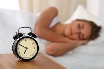 Beautiful young woman sleeping and smiling while lying in bed comfortably and blissfully on the background of alarm clock is going to ring. Sunbeam dawn  on her face.