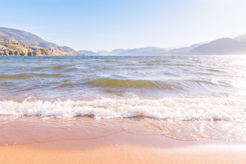 Waves crashing onto sandy beach with mountains, sunshine, and blue sky in background