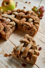 A piece of the homemade apple pie and brown raisins on light wooden background. The applepie decorated with fresh apples and grapes on blurred background. Horizontal image. 