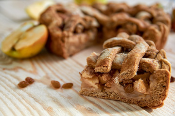 Obraz na płótnie Canvas A piece of the homemade apple pie with brown raisins close up on light wooden background. The apple pie decorated with fresh apples and raisins on blurred background. 