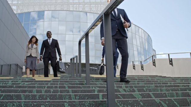 Tracking of business people walking on stairs in front of modern office building; young woman talking with African colleague and two men greeting each other with handshake