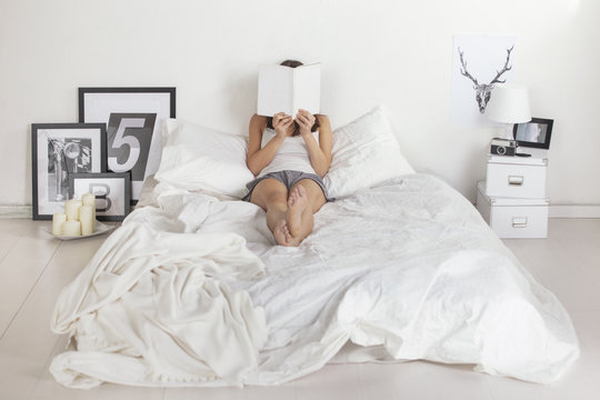 Woman reading with the face covered by the book. Relaxation on the white bedroom.