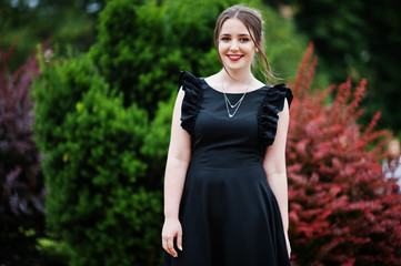 Portrait of a gorgeous young girl in black dress walking on the pavement in the park on a prom day. - obrazy, fototapety, plakaty