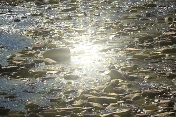 stones in a shallow river and a reflection of the sun in the water at the center of the frame