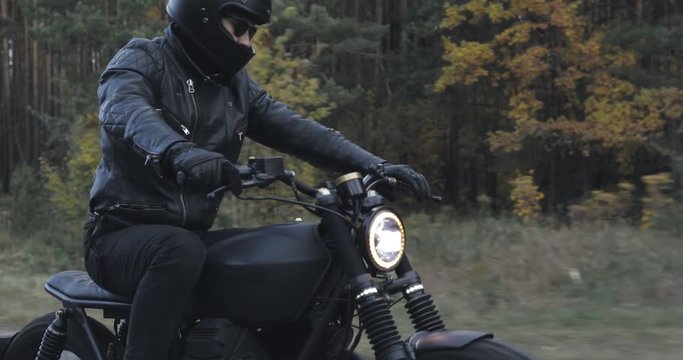 A guy in a black leather jacket and helmet riding a classic motorcycle on a forest road.