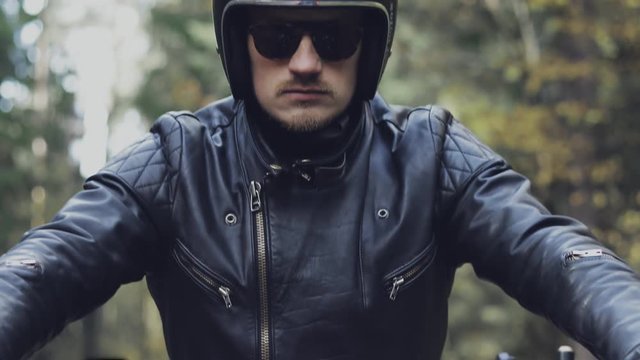 A guy in a black leather jacket and helmet riding a classic motorcycle on a forest road.