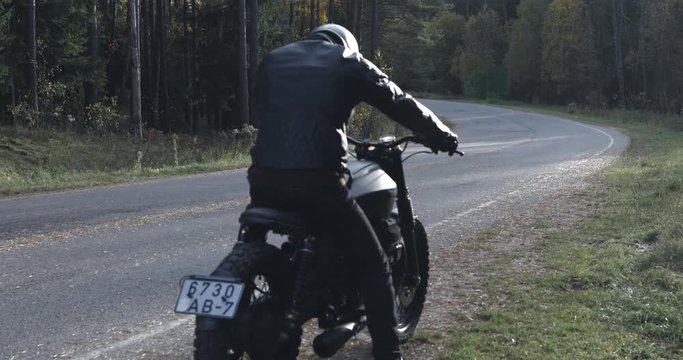 A guy in a black leather jacket and helmet riding a classic motorcycle on a forest road. View from the back