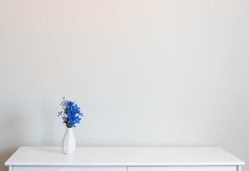 Small white vase of blue delphinium flowers on cabinet against neutral background with copy space