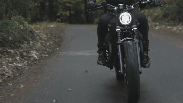 A guy in a black leather jacket and helmet riding a classic motorcycle on a forest road.
