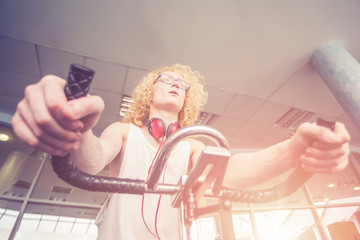 Curly blond man with glasses and red headphones training on a exercise bike in the gym in the...