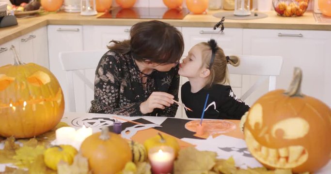 Mother and daughter sitting at table near jack-o-lantern, making Halloween decorations and kissing each other.
