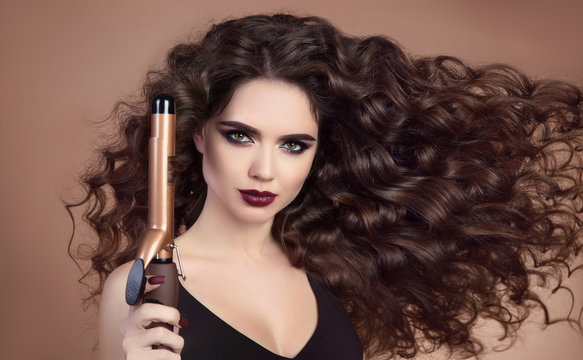 Salon Beauty. Curly hair. Close-up of brunette girl with blowing hairstyle holding Professional curling iron. Marsala eye shadow makeup. Glamour female isolated on beige background.