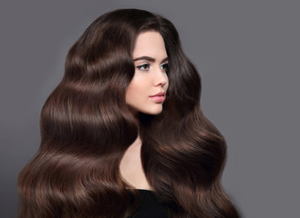 Naklejka premium Healthy long wavy hair. Brunette girl beauty portrait with shiny brown hair. Beautiful model with hairstyle and makeup isolated on studio grey background. Shampoo care product.