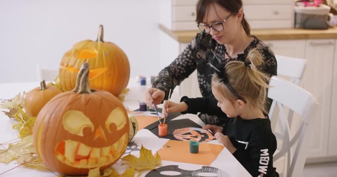 Woman in glasses and her daughter in Halloween costume sitting at table and painting jack-o-lantern made from paper.