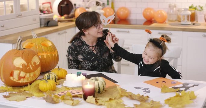 Woman in glasses sitting at kitchen table and looking at her daughter fitting white mask for Halloween party.