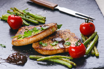 Grilled pork chops, steaks with vegetables, tomatoes, beans and sauce on a black slate. Fresh meat with foam. Dark background. Grilled and barbecue concept. 