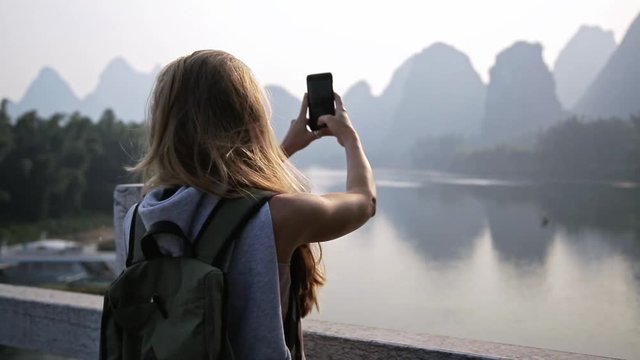 Young blonde woman is taking photo with smartphone and looking at panorama view of Li river in Yangshuo, Guilin, China. Travel, destination and adventure concept.