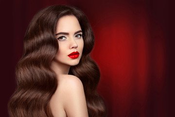 Fototapeta premium Beauty hair. Brunette girl portrait with red lips makeup and long shiny wavy hair. Beautiful model with healthy hairstyle isolated on drapery curtains dark background. Gorgeous Glamour Lady.