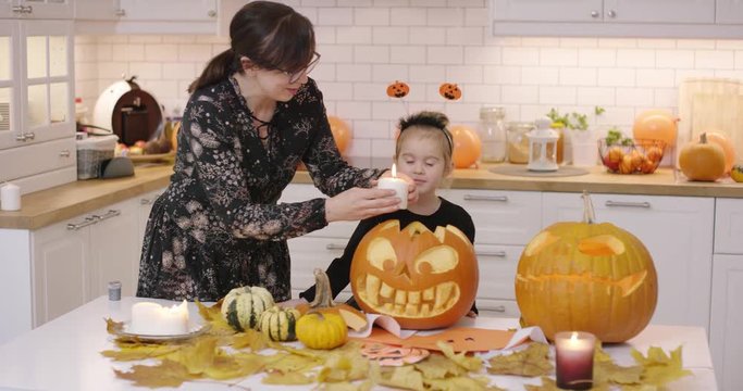 Woman in glasses standing in kitchen near her little daughter and putting big candle into creepy jack-o-lantern.