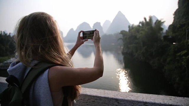 Young blonde woman is taking photo with smartphone and looking at panorama view of Li river in Yangshuo, Guilin, China. Travel, destination and adventure concept.