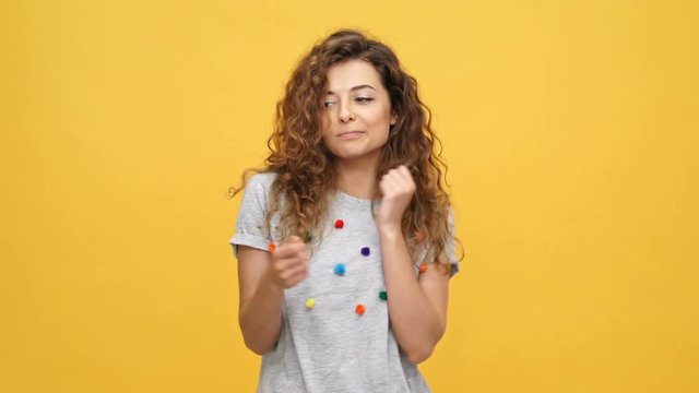 Happy funny curly woman in t-shirt rejoice and dancing while looking at the camera over yellow background