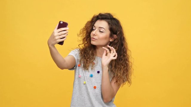 Smiling happy curly woman in t-shirt making selfie on smartphone over yellow background
