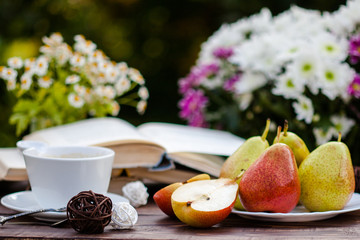 Summer romantic still life. Fresh ripe organic Pears, bouquet of field daisy and chrysanthemum flowers, cup of coffee, book on wooden table surface. Natural trees background. Fruit healthy lunch