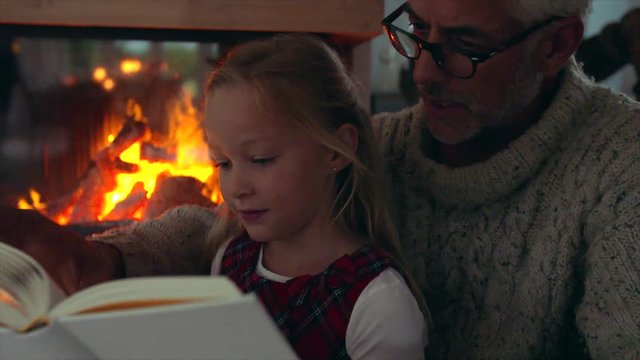 Little girl reading book with grandfather near fireplace at home. Senior man with grandaughter reading a book.