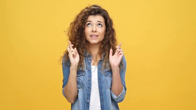 Young curly woman in denim jacket praying with crossed fingers and hands in pray gesture over yellow background