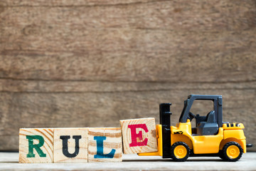 Yellow plastic toy forklift hold letter block E to complete word rule on wood background