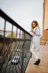 Stylish curly blonde model girl wear on white with cup of coffee and mobile phone at hand posing against railings.
