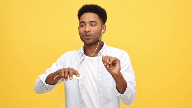 Happy african man in shirt listening music by headphones and dancing while sings and looking at the camera over yellow background