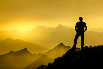 Man standing on summit enjoying sunrise and spectacular layered mountain range silhouettes.