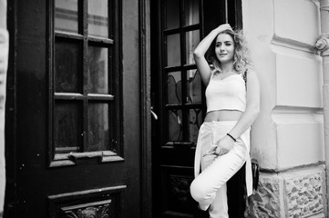 Stylish curly blonde model girl wear on white posing against old wooden door.