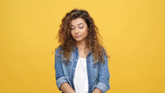 Bored curly woman in denim shirt touching face and looking around over yellow background