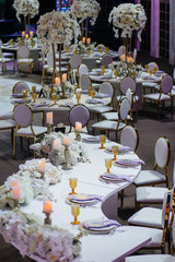 Long dinner table with white flower garlands, shiny candles on silver candleholders, golden glasses and violet cloth