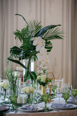 Tall vase with large green leaves and orchids stands in the middle of dinner table