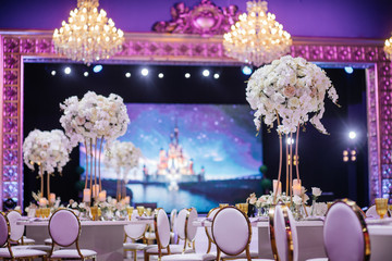 Large white bouquets of roses stand on dinner tables decorated with golden glasses and violet cloth