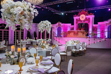 Large white bouquets of roses stand on dinner tables decorated with golden glasses and violet cloth