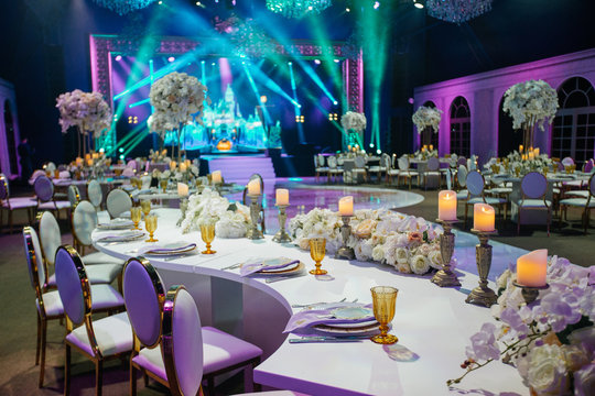 Long dinner table decorated with white flowers, shiny candles and golden glasses stands in a beautiful hall
