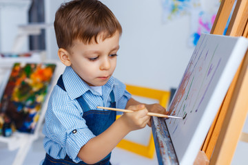 Cute, serious and focused, three years old boy in blue shirt and jeans apron painting on canvas...