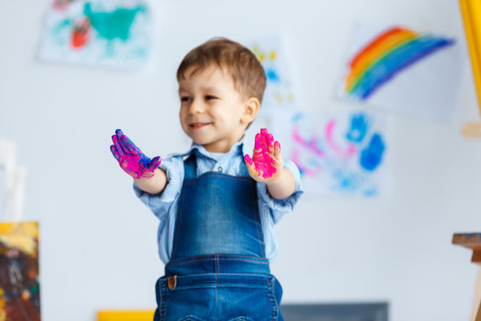 Cute, happy, white, three year boy in blue shirt and jeans showing his dirty hands in watercolors. Little child having fun after painting. Concept of early childhood education, happy family, parenting