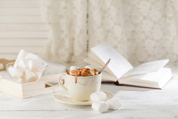 Mug of hot chocolate with marshmallows, on light wooden background with open book