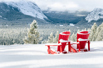 Snow Covered Red Adirondack Chairs Facing a Frozen Forested Valley on a Snowy Winter Day. Banff...