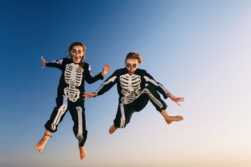 Obraz premium Two young girls in scary skeleton costumes jump high in air with fun before Halloween night party on sunset sea beach. Active people, lifestyles and funny event celebrations on holidays