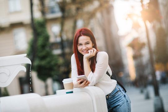 Young Woman Standing At The Street Next To Drinking Fountain Using Mobile Phone And Drinking Coffee To Go  