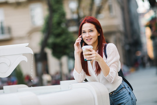 Young woman standing at the street next to drinking fountain talking on mobile phone and drinking coffee to go  
