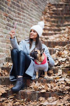 Woman enjoying outdoors with her adorable French bulldog. They sitting on park stairs covered with withered autumn leaves. 